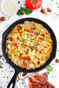 A cast iron skillet filled with a baked dish topped with melted cheese, bacon, tomato, and greens, surrounded by bacon strips and fresh vegetables on a white surface.