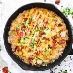 A cast iron skillet filled with a baked cheesy dish topped with chopped lettuce, tomato, and bacon, surrounded by fresh ingredients and crispy bacon strips on the side.