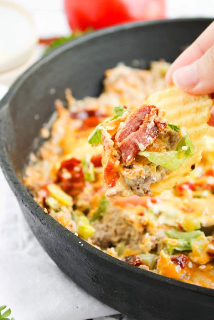 A hand holds a potato chip topped with a cheesy, bacon-loaded dip, lifted from a cast iron skillet filled with the same dip.