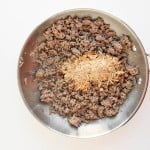 A stainless steel pan with cooked ground beef and a mound of dry seasoning mix on top, set on a white surface.