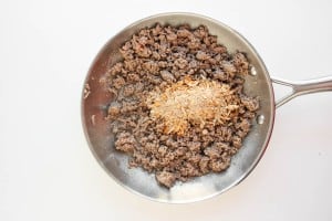 A stainless steel pan with cooked ground beef and a mound of dry seasoning mix on top, set on a white surface.