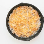 A cast iron skillet filled with an uncooked mixture topped with a layer of shredded cheese on a white background.