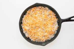 A cast iron skillet filled with an uncooked mixture topped with a layer of shredded cheese on a white background.