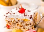 A close-up of a slice of cake with white frosting, chocolate chips, a maraschino cherry on top, and visible fruit and chocolate pieces inside.