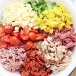 A white bowl filled with diced lettuce, mozzarella cheese, banana peppers, grape tomatoes, ham, salami, turkey, and pepperoni&mdash;each ingredient arranged in sections for a classic Grinder Chopped Salad. Wooden salad utensils rest beside the bowl.