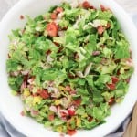 A white bowl filled with Grinder Chopped Salad&mdash;lettuce, tomatoes, meats, and cheese&mdash;sits next to a striped cloth and wooden salad utensils.