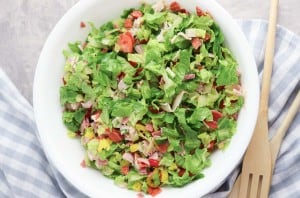 A white bowl filled with Grinder Chopped Salad—lettuce, tomatoes, meats, and cheese—sits next to a striped cloth and wooden salad utensils.