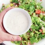 A hand holds a small bowl of creamy salad dressing above a large bowl of Grinder Chopped Salad, brimming with lettuce, tomatoes, and other fresh vegetables.