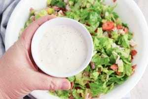A hand holds a small bowl of creamy salad dressing above a large bowl of Grinder Chopped Salad, brimming with lettuce, tomatoes, and other fresh vegetables.