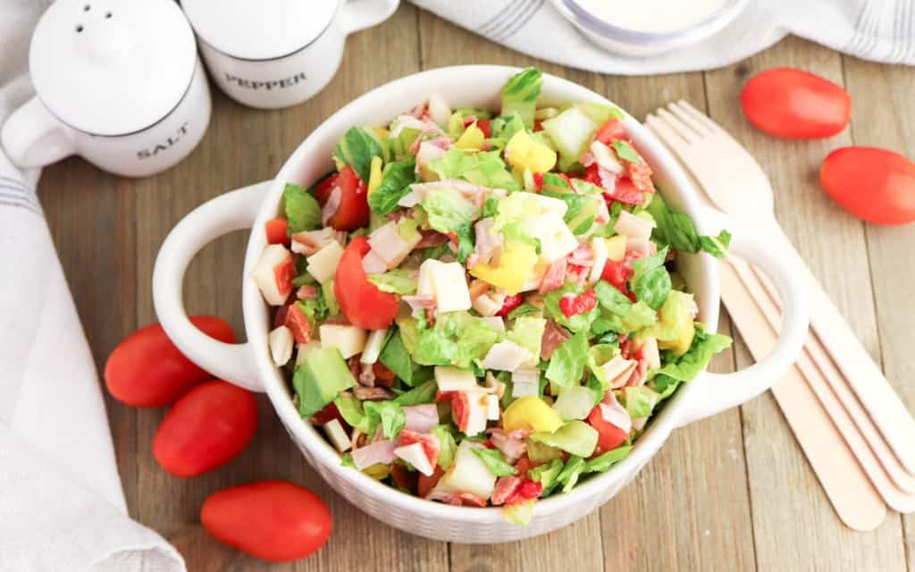 A bowl of Grinder Chopped Salad with lettuce, tomatoes, cheese, and veggies sits on a wooden table, surrounded by grape tomatoes, a salt and pepper grinder set, and wooden utensils.
