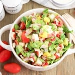 A bowl of Grinder Chopped Salad with lettuce, tomatoes, cheese, and veggies sits on a wooden table, surrounded by grape tomatoes, a salt and pepper grinder set, and wooden utensils.