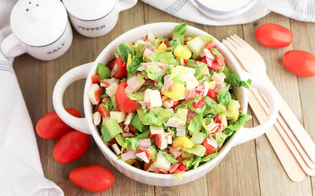 A bowl of Grinder Chopped Salad with lettuce, tomatoes, cheese, and veggies sits on a wooden table, surrounded by grape tomatoes, a salt and pepper grinder set, and wooden utensils.