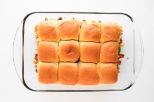 A glass baking dish containing twelve baked slider sandwiches arranged in a 3x4 grid, photographed from above.