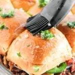 A basting brush applies a herb and butter mixture onto a tray of assembled sandwich sliders filled with meat and vegetables.