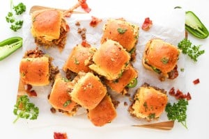 A tray of small sandwiches filled with meat and topped with herbs, arranged on parchment paper with parsley and jalapeño slices as garnish.