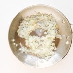 Diced onions cooking in a stainless steel pan on a white surface.