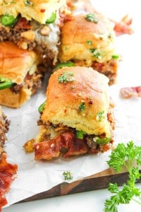 A close-up of a small sandwich with a golden bun, ground beef, bacon, melted cheese, jalapeños, and parsley on parchment paper.