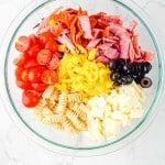 A glass bowl contains Grinder Pasta Salad ingredients: cherry tomatoes, sliced pepperoni, salami, pepperoncini, black olives, rotini pasta, and cubed mozzarella arranged in separate sections.