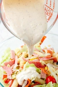 Creamy dressing is being poured from a measuring cup onto a bowl of Grinder Pasta Salad with lettuce, sliced meats, and cherry tomatoes.