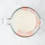 A glass measuring cup filled with creamy white sauce for Grinder Pasta Salad, placed on a white marble surface.