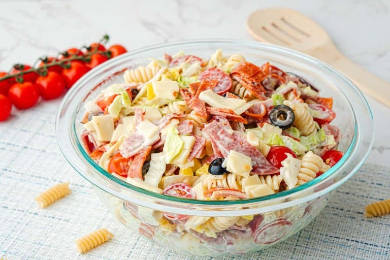 A glass bowl filled with Grinder Pasta Salad—rotini pasta, lettuce, tomatoes, olives, cheese, and sliced meats—sits on a placemat next to cherry tomatoes and a wooden spoon.