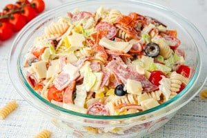 A glass bowl filled with Grinder Pasta Salad featuring rotini pasta, lettuce, tomatoes, olives, cheese cubes, and slices of cured meat, all tossed together with a creamy dressing.