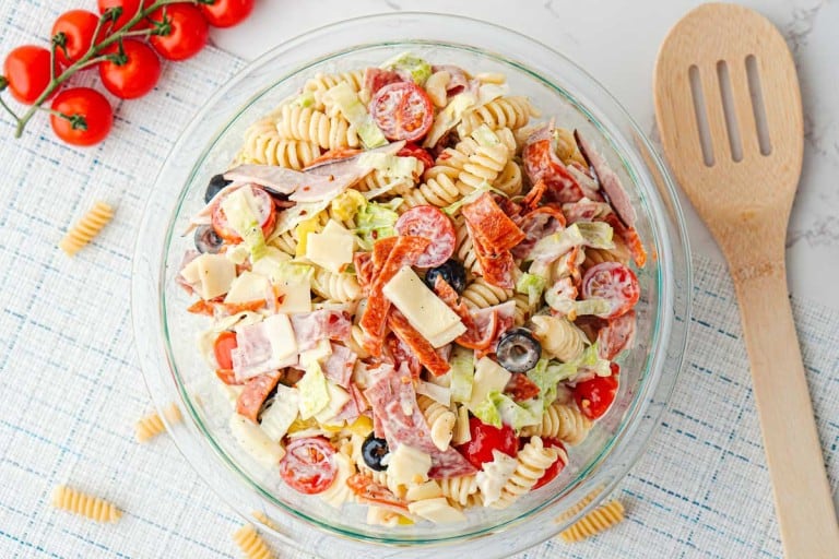 A glass bowl of Grinder Pasta Salad with rotini noodles, sliced cherry tomatoes, olives, deli meats, cheese, and lettuce sits on a light placemat next to a wooden spoon and vine tomatoes.