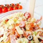 A glass bowl of Grinder Pasta Salad with rotini, sliced salami, cheese, black olives, lettuce, and cherry tomatoes. A wooden spoon rests in the bowl; fresh cherry tomatoes are scattered in the background.