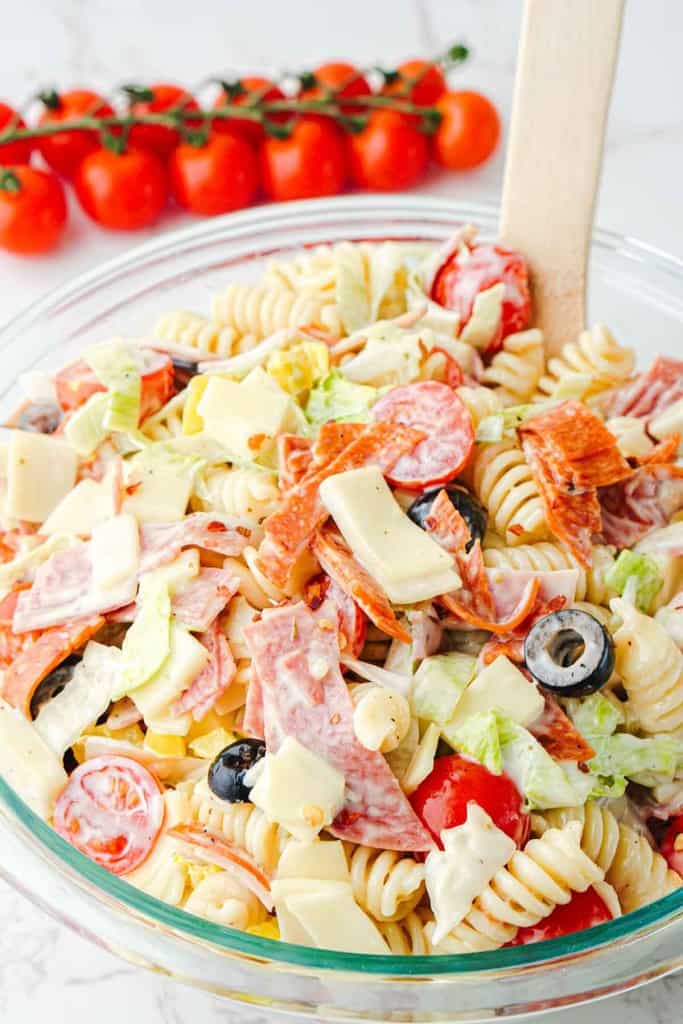 A glass bowl filled with Grinder Pasta Salad featuring rotini, cheese, lettuce, olives, cherry tomatoes, and sliced meats, with a wooden spoon; vine tomatoes in the background.