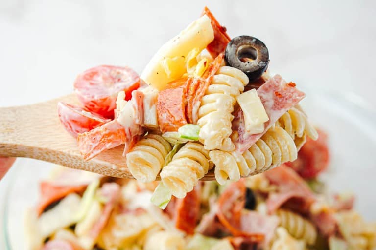 A wooden spoon holds a serving of Grinder Pasta Salad featuring rotini, salami, cheese, olives, lettuce, and tomatoes tossed in a creamy dressing.