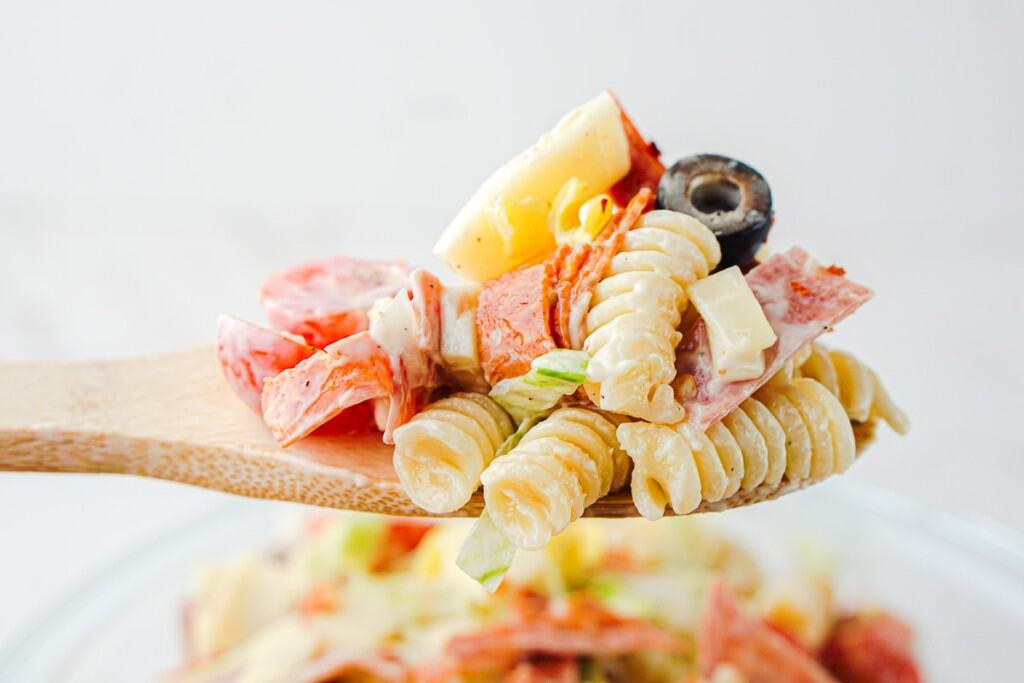 A wooden spoon holds a serving of Grinder Pasta Salad featuring rotini, cheese cubes, salami, black olive slices, and fresh vegetables.