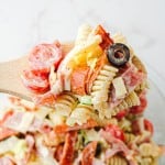 A wooden spoon holds Grinder Pasta Salad with rotini, cherry tomatoes, cheese cubes, pepperoni, olives, and diced vegetables over a glass bowl.