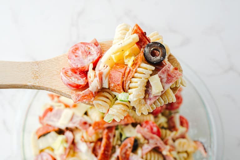 A wooden spoon holds Grinder Pasta Salad with rotini, cherry tomatoes, cheese cubes, pepperoni, olives, and diced vegetables over a glass bowl.