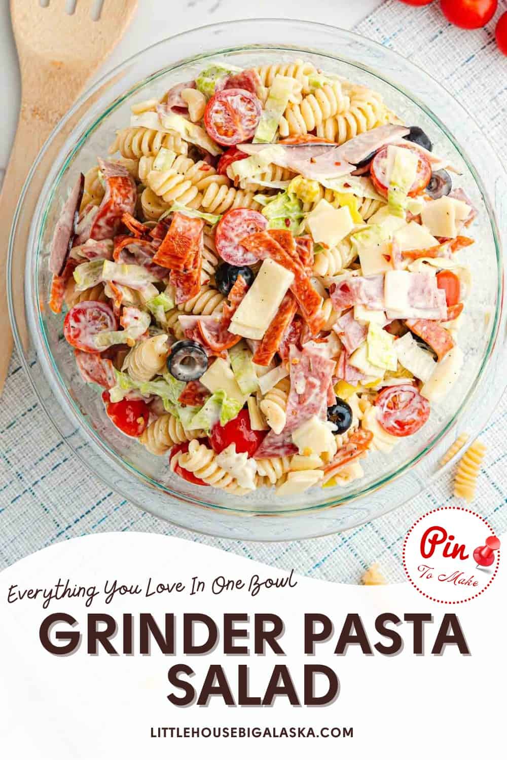 A glass bowl filled with grinder pasta salad made of rotini pasta, sliced deli meats, cheese, cherry tomatoes, olives, and lettuce, on a white surface with text below.