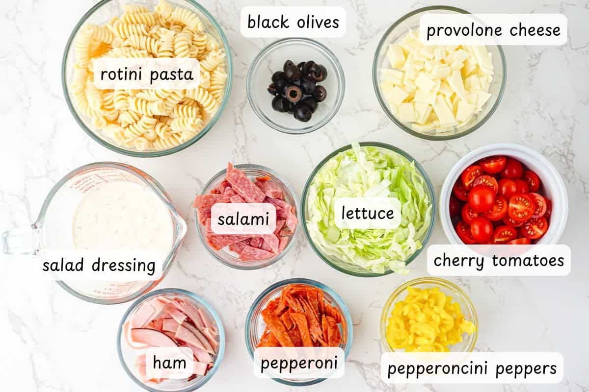 Nine bowls on a white surface contain rotini pasta, black olives, provolone cheese, salad dressing, salami, lettuce, cherry tomatoes, ham, pepperoni, and pepperoncini peppers.