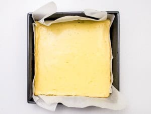 A square cheesecake in a parchment-lined baking pan, viewed from above, with a smooth, pale yellow top.