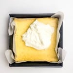 A square baking pan lined with parchment paper holds a baked yellow cake topped with a dollop of whipped cream, viewed from above on a white background.