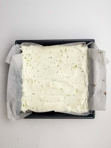 A rectangular baking pan lined with parchment paper holds a dessert topped with a layer of white whipped cream and sprinkled green zest.