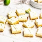 Twelve key lime pie bars with a crumb crust and whipped topping are arranged on parchment paper, with lime zest sprinkled on top and fresh lime wedges nearby.
