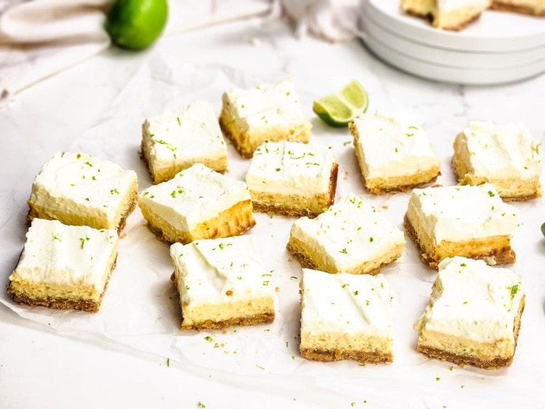 Twelve key lime pie bars with a crumb crust and whipped topping are arranged on parchment paper, with lime zest sprinkled on top and fresh lime wedges nearby.