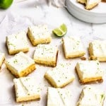 Nine frosted dessert bars are arranged on parchment paper, garnished with lime zest. A cut lime and additional bars on a plate are visible in the background.