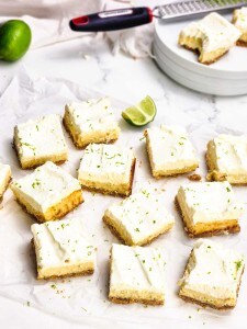Nine frosted dessert bars are arranged on parchment paper, garnished with lime zest. A cut lime and additional bars on a plate are visible in the background.