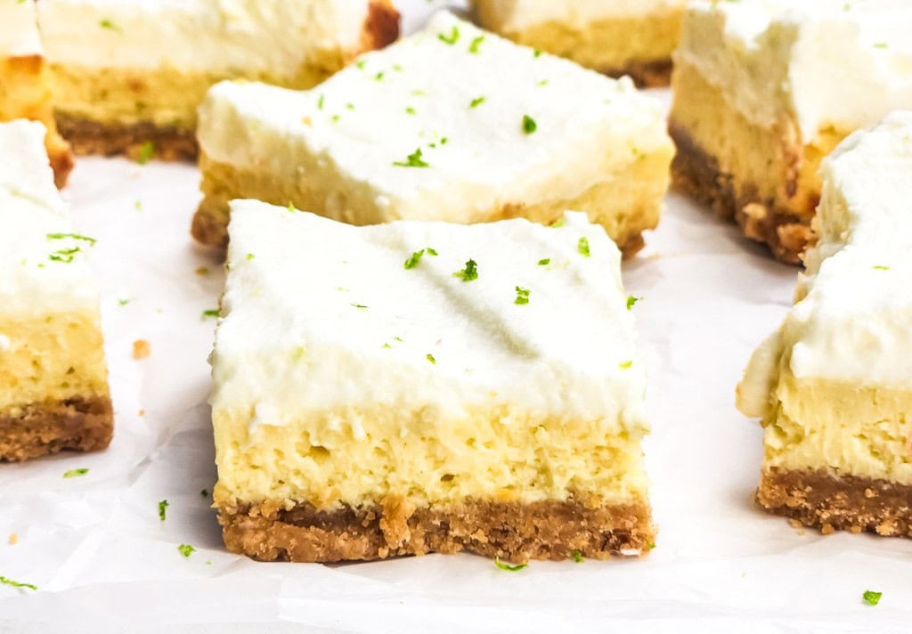Close-up of several lime cheesecake bars with a crumbly crust, creamy filling, and whipped topping, garnished with lime zest, arranged on white parchment paper.