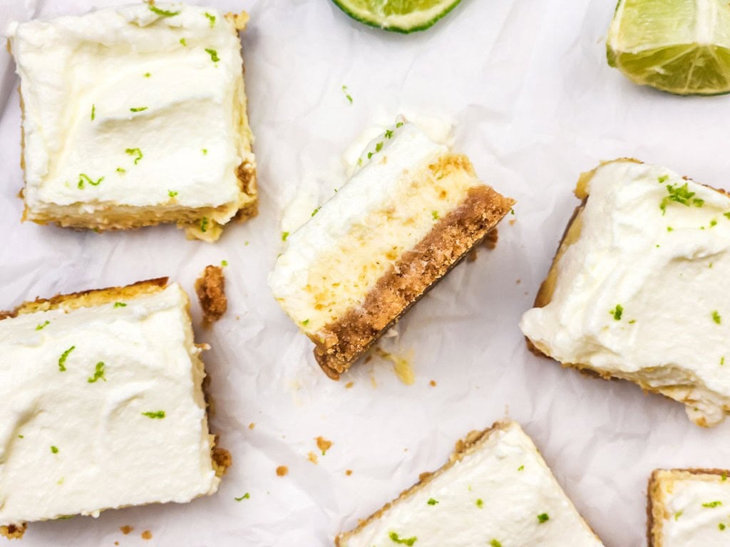 Several key lime pie bars with graham cracker crust and whipped cream topping are arranged on parchment paper, with lime slices nearby.