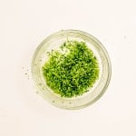 A clear glass bowl filled with green lime zest, placed on a plain white surface.