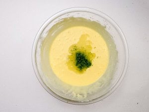 A glass bowl containing a yellow batter topped with green zest and a small amount of liquid, placed on a white surface.
