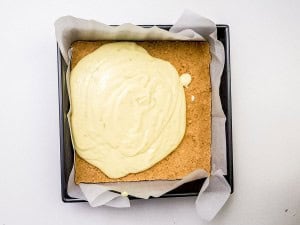 A square baking pan lined with parchment paper contains a baked crust with a layer of yellow batter spread on top, ready to be further baked.