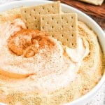 A white bowl filled with creamy dip topped with ground spices and crumbs, garnished with two rectangular crackers. More crackers are visible on a plate in the background.