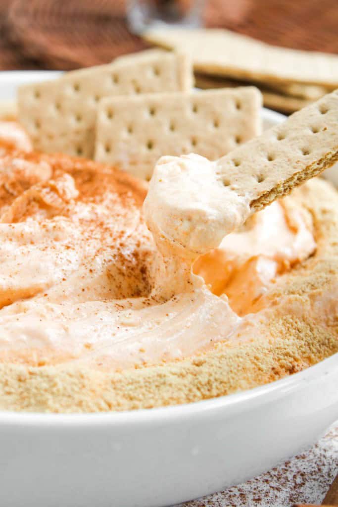 A rectangular cracker is dipped into a creamy, orange-tinted dip in a white bowl, with more crackers and a dusting of spice on top.