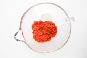 A clear glass mixing bowl contains a block of cream cheese and a mound of tomato paste, placed on a white surface.